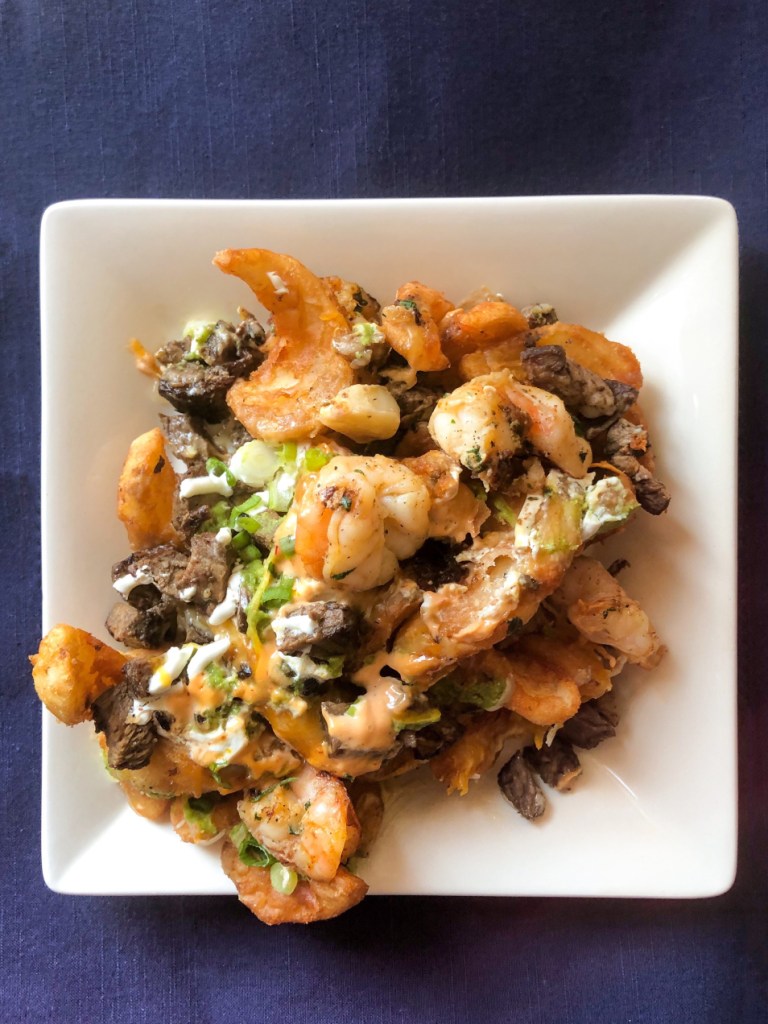 A plate of Little Miner Taco’s Carne Asada Fries. It's topped shrimp, cheese, green onion, chives, sour cream, and sriracha aioli. Little Miner Taco is a restaurant with multiple locations in the Washington, D.C. Metro Area.