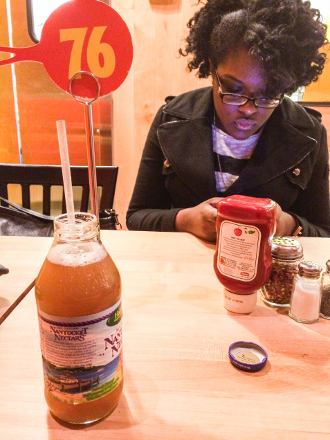 A woman waiting for her food to arrive to her table; apple juice and condiments are on the table.