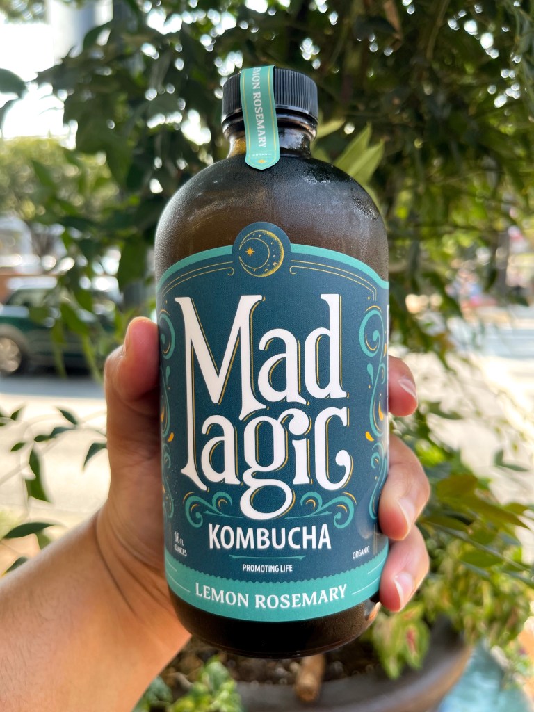 A hand holding a bottle of Lemon Rosemary kombucha from Mad Magic, a local kombucha crafted in Virginia.