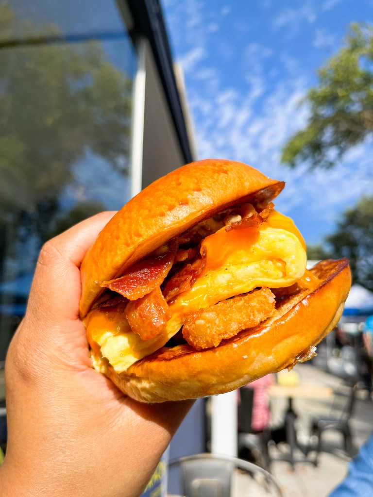 A hand holding a sandwich called "The Animal." It contains bacon, sausage, scrambled egg, tater tots, and cheese. This sandwich is from Cracked Eggery, which is located in Washington, D.C.