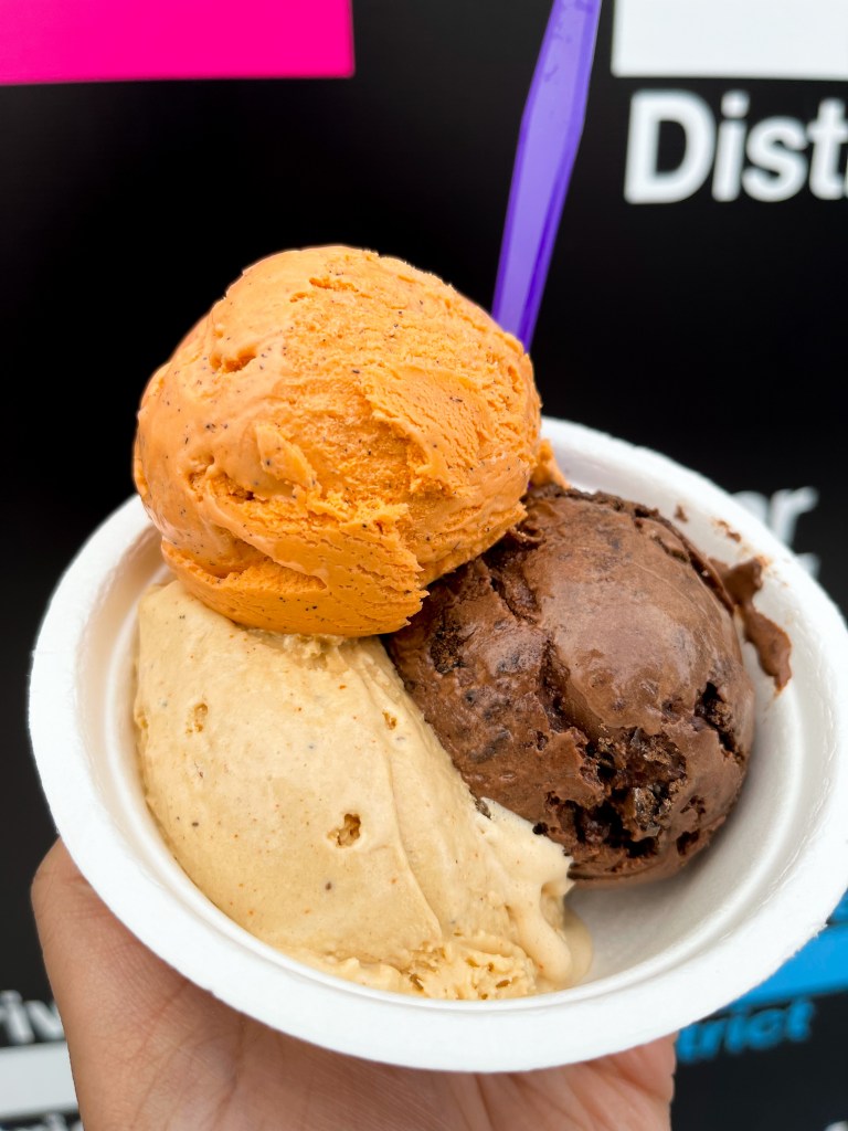 A hand holding a bowl of ice cream from the Charmery in Columbia, MD. The flavors pictured are Thai Tea, Old Bay Caramel, Choccy Peppy Cookie.